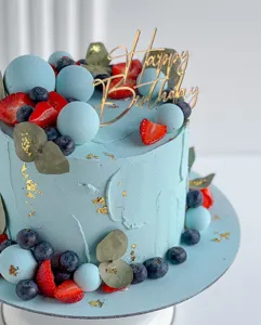 Gros plan d'un gâteau d'anniversaire bleu avec cake topper acrylique doré et détails fruits.