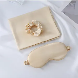 Silk sleep set detail showing champagne pillowcase, eye mask and scrunchie on white fabric.