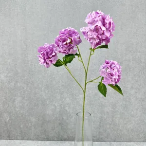 Nahaufnahme künstlicher violetter Hortensienblüten mit grünen Blättern in klarer Glasvase.