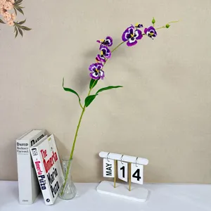Artificial purple pansy orchid flower stem in glass vase with books and calendar decor.