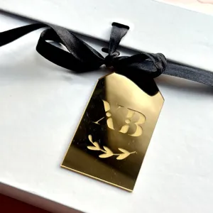 Close-up detail of gold mirror acrylic pendant tag with black ribbon on white box packaging.