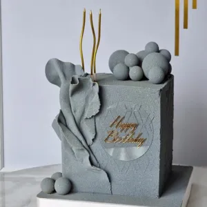 Minimalist grey square birthday cake decoration with gold candles and geometric sphere accents.