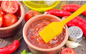 Detail shot of a yellow silicone basting brush dipping into sauce with kitchen props.