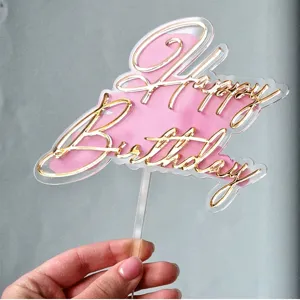 Vue de detail de cake topper dore en acrylique Joyeux Anniversaire avec fond rose fete.
