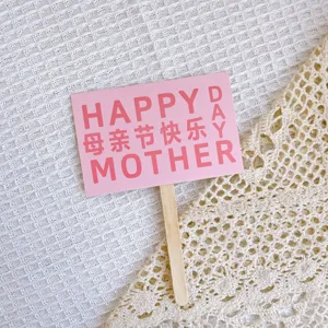 Close-up detail of a pink Mother's Day cake topper card with wooden stick on textured fabric.