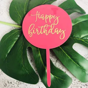 Rosa runder Acryl-Kuchentortenaufsatz mit goldenem Happy Birthday Text auf grünem Monstera Blatt.
