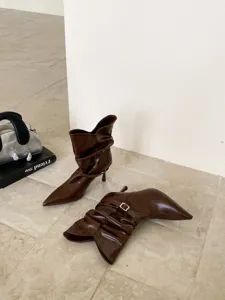 Detalle de botas marrones de punta fina con tacón alto, hebilla elegante y diseño fruncido único.