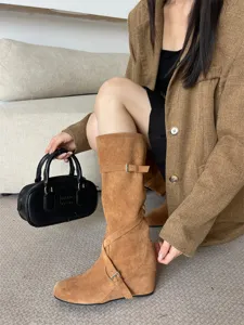 Primer plano detallado de botas altas elegantes de ante marrón con hebilla y tacón de cuña.
