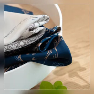 Detail shot of folded silk scarf in white bowl on wooden table, elegant patterned fabric.