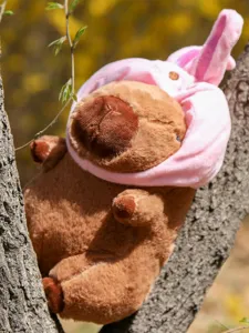 Süßes Kapibara Plüschtier mit rosa Hasenmütze, flauschige Stoffpuppe im Freien am Baumast dekoriert.