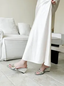 Close-up of stylish silver metallic open-toe wedge sandals paired with white skirt in room.
