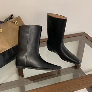 Detailed shot of black leather square toe flat boots on glass display table with luxury bag prop.