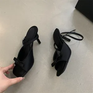 Nahaufnahme von schwarzen Satin-High-Heel-Sandalen mit Schleife und Knöchelriemen-Detail.