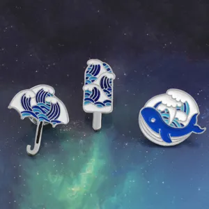 Primo piano dettagliato di spille smaltate con onde blu e bianche, balena, ombrello e gelato.