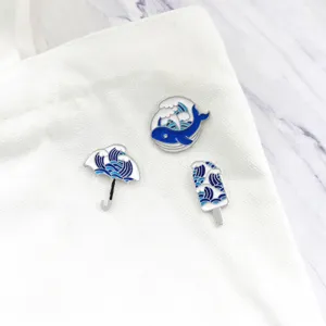 Primo piano della spilla in smalto con onde blu e bianche, con spille balena e ombrello.