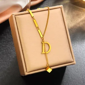 Pendentif de collier lettre D en acier inoxydable plaqué or 18k sur boîte à bijoux beige.