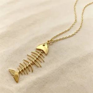 Close up of a delicate gold fish skeleton pendant necklace resting on textured sand background.