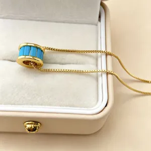 Close-up detail van een goudkleurige cilindrische hangketting met blauwe inzet in doos.