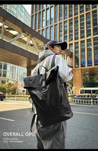 Outdoor detail shot of a black PU leather roll-top backpack worn by a stylish male model.