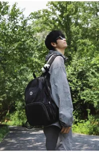 Outdoor detail view showing the black backpack worn by a model in a natural green park setting.