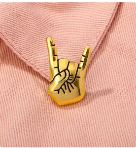 Close-up detail shot of a gold-plated rock hand gesture metal brooch pin on pink fabric texture.