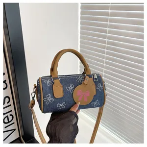 Denim barrel handbag detail showing white bow print, brown leather handle and zipper.