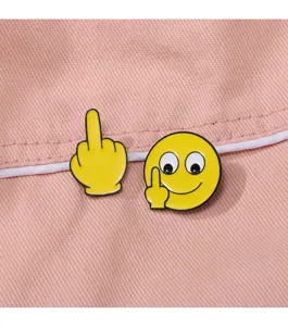 Primo piano dettagliato della spilla smaltata con emoji e dito medio su tessuto rosa.