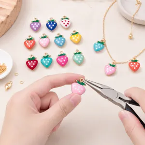 Detailopname van kleurrijke geëmailleerde aardbei-bedels met gouden ringetjes en een tang.