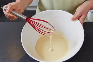 Detailaufnahme eines manuellen Silikon-Schwingbesens mit Edelstahlgriff beim Rühren im Teig.