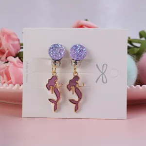 Close-up detail of cute purple glitter and mermaid clip-on earrings on a white card display.