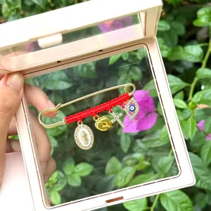Gold safety pin brooch with red braided cord, evil eye and cross charms for fashion styling.