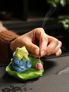 Close-up detail of ceramic frog incense burner on lotus leaf with burning incense stick.