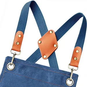Blue canvas apron detail showing cross back straps, brown leather patches, and durable metal hardware.