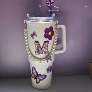 Primo piano del bicchiere bianco decorato con catena di perle, lettera M e patch di farfalle.