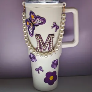 Dettaglio bicchiere termico bianco con toppe ricamate farfalla e fiore viola e lettera M.