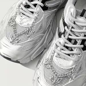 Close-up detail shot of silver chunky sneakers decorated with metallic bat pendant chains.