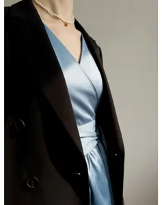 Élégante robe portefeuille en satin bleu associée à un blazer noir et un collier de perles.