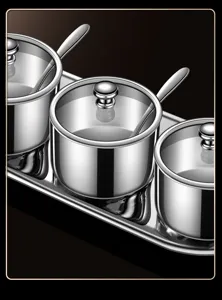 Stainless steel condiment jar set with glass lids, spoons, and tray for kitchen storage.
