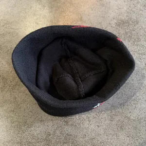 Close-up detail view of a black knitted beanie hat, showing textured fabric and knit construction.