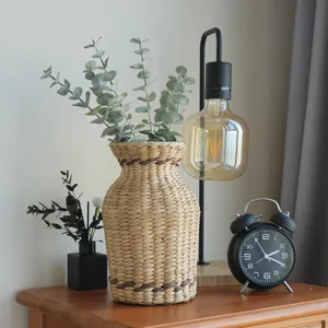 Nahaufnahme einer geflochtenen Strohvase auf einem Holztisch mit dekorativen Trockenpflanzen.