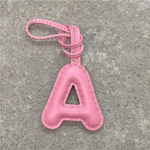 Gros plan détail d'un charm de sac lettre A en cuir rose gonflé avec une lanière blanche.