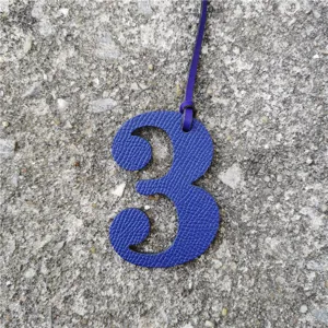 Close-up detail view of a blue textured leather number 3 bag charm with hanging strap.