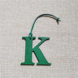 Gros plan du détail d'un charm de sac en cuir vert en forme de lettre K avec une lanière.