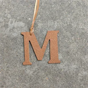 Close-up detail of luxury leather M letter charm pendant, textured tan leather material.
