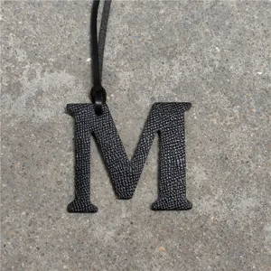 Detailed close-up of a black leather letter M bag charm with strap on textured surface.