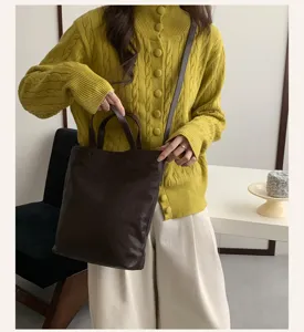 Detail view of minimalist brown leather tote bag held by model wearing knit sweater in room.