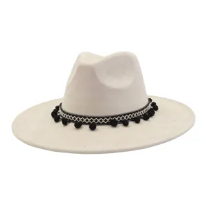 Detail view of beige suede fedora hat with black pom-pom band and wide brim design.