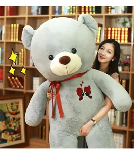 Large grey plush teddy bear with red ribbon and heart embroidery, held by model in room.
