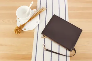 Close-up of a dark brown leather clutch pouch with wrist strap and zipper on a striped runner.