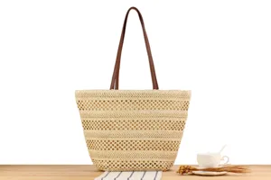 Detail shot of a large straw tote bag with brown leather handles displayed on a wooden table.